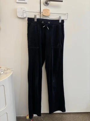 Svarta mjukisbyxor från juciy couture med snörning - Svarta mjukisbyxor i mjukt sammetstyg med snörning i midjan och praktiska fickor både fram och bak. Baktill finns två fickor med lock och knappar. Byxorna har en rak passform och känns riktigt sköna att ha på sig. Dom är från juciy couture och köptes för 800kr 