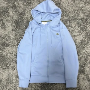 Ljusblå hoodie från Lacoste - Snygg ljusblå hoodie från Lacoste i klassisk passform. Tröjan har huva med snörning, dragkedja framtill och två fickor. Ikoniska Lacoste-krokodilen är broderad på bröstet. Perfekt för chill dagar och enkel att matcha med jeans eller joggers.