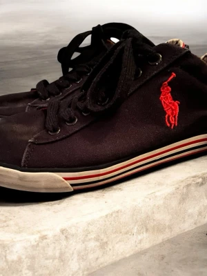 Svarta sneakers från Polo Ralph Lauren - Svarta sneakers från Polo Ralph Lauren med röd broderad logga på sidan och snörning. Yttermaterialet är bomullstyg och sulan är vit med röda och svarta ränder samt röd undersida. Klassisk rund tå och platt sula. Märkesdetaljer både på insidan och undersidan av skon.