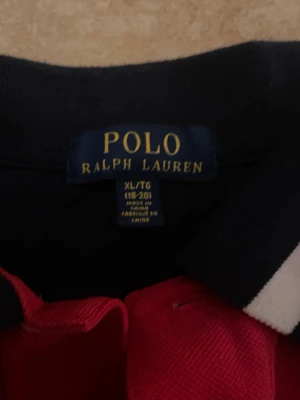 Röd och marinblå pikétröja Polo Ralph Lauren - Säljer en snygg pikétröja från Polo Ralph Lauren i storlek XL. Tröjan är röd med marinblå detaljer och har en vit broderad logga på bröstet samt ett broderat emblem med siffran 3. Klassisk krage med vit och marinblå rand, korta ärmar och knappar framtill. PASSAR ÄVEN M OCH S!!    vid frågor skriv till mig gärna!