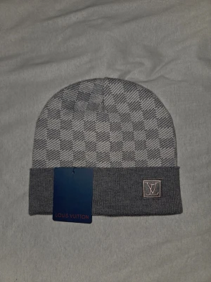 Grå rutig mössa från Louis Vuitton - Säljer en snygg grå mössa från Louis Vuitton med klassiskt rutigt mönster och vikt kant. Diskret LV-logga framtill och exklusiv känsla. Perfekt accessoar för dig som vill ha något stilrent och lyxigt till vintern.