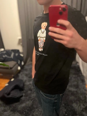 Svart Polo Bear t-shirt från Ralph Lauren - Säljer en svart t-shirt från Ralph Lauren med ikoniskt Polo Bear-tryck på bröstet. Björnen har en vit tröja med färgglada detaljer och rutiga byxor. T-shirten är kortärmad och har en klassisk passform, perfekt för dig som gillar streetwear med lite extra attityd.