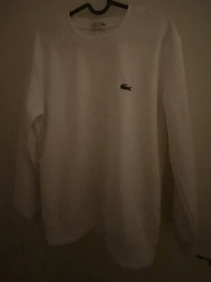 Vit sweatshirt - Säljer en clean vit sweatshirt från Lacoste med klassisk krokodillogga broderad på bröstet. Tröjan har rund hals, långa ärmar och ribbade muddar vid ärmslut och nederkant. Regular fit och mjukt material som är skönt mot huden. (Obs vet ej om äkta eller inte)