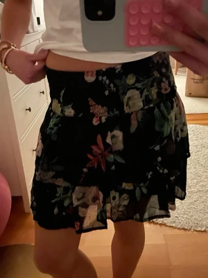 Svart blommig kortkjol med volang - Säljer en svart kortkjol med blommigt mönster och volangdetaljer. Skitsnygg till sommaren! Mycket bra skick, inga tecken på användning. Storlek 146/152 men passar som xs! 