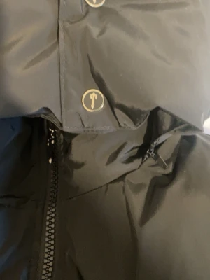 Svart pufferjacka från Trapstar storlek S - Svart pufferjacka från Trapstar med huva och fet logga broderad på ryggen. Jackan har dragkedja framtill, stora fickor och Trapstar-märke på bröstet. Perfekt för kalla dagar och har en riktigt clean look.