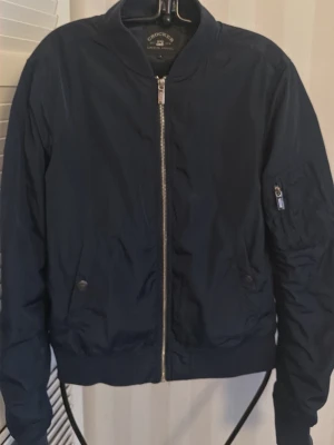 Mörkblå bomberjacka från Crocker - Snygg mörkblå bomberjacka från Crocker med klassisk silvrig dragkedja framtill och på ärmen. Jackan har ribbade muddar, två fickor med knapp och en liten ficka med dragkedja på ärmen. Perfekt för dig som gillar stilren och enkel design.