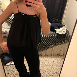 Svart linnetopp med pärldetaljer - Säljer en svart, luftig linnetopp med tunna axelband och pärldetaljer längs den rynkade halsringningen. Toppen har en loose passform och är perfekt för varma dagar. Materialet känns lätt och svalt mot huden.