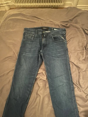 Replay Anbass jeans blå storlek W31 L30 - Säljer ett par snygga Replay Anbass jeans i mörkblå tvätt med klassisk femficksdesign och dragkedja. Jeansen har en slim passform och är tillverkade i mjukt jeansmaterial med diskreta slitningar. Perfekt för dig som gillar stilrena och moderna jeans.