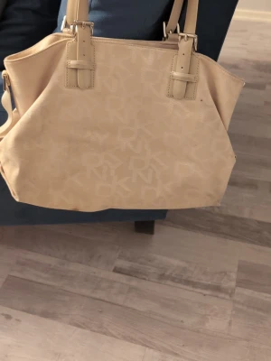 Beige axelväska från DKNY - Säljer en beige axelväska från DKNY med snyggt monogrammönster och silverfärgade detaljer. Väskan har både handtag och justerbar axelrem, samt praktisk dragkedja och innerfack. Perfekt storlek för att få plats med allt du behöver.