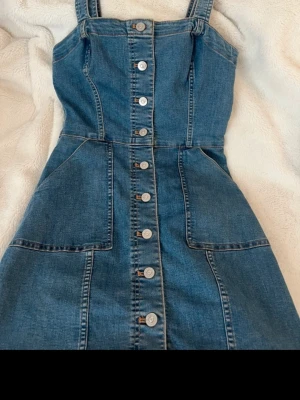 Jeansklänning med knappar från H&M Divided - Säljer en snygg jeansklänning från H&M Divided i storlek 34. Klänningen har breda axelband, knappar hela vägen framtill och markerad midja. Klassisk blå denim med coola sömdetaljer och fickor framtill. Perfekt att styla med sneakers eller boots.