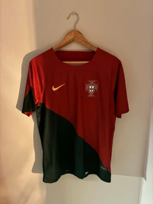 Portugal Nike fotbollströja röd/grön S - Portugal fotbollströja från Nike i rött och mörkgrönt med diagonal färgdelning. Broderat landslagsemblem på bröstet och gul Nike-logga. Tillverkad i Dri-FIT polyester för bra andningsförmåga. Perfekt för dig som älskar fotboll och vill bära landslagets färger.