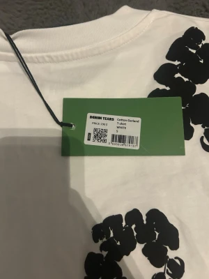 Vit Cotton Garland t-shirt Denim Tears - Säljer en vit t-shirt från Denim Tears med svarta blommiga tryck över hela tröjan. Modellen är klassisk med rund hals och korta ärmar. Tillverkad i mjuk bomull som känns skön mot huden. Perfekt för dig som gillar streetwear och unika prints.