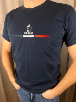 Mörkblå Moncler t-shirt med tryck - Snygg mörkblå t-shirt från Moncler med klassiskt tryck på bröstet i vitt, rött och marinblått. T-shirten är kortärmad och har rund hals. Tillverkad i mjuk bomull som känns skön mot huden. Perfekt för dig som gillar stilrena och exklusiva plagg.