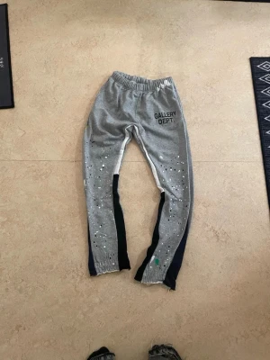 Grå mjukisbyxor från Gallery Dept. - Säljer ett par grå mjukisbyxor från Gallery Dept. med elastisk midja och coolt stänkmönster i vitt och svart på benen. Byxorna har svarta och vita paneler längs sidorna och råa kanter vid bensluten. Perfekt för en avslappnad och trendig look.