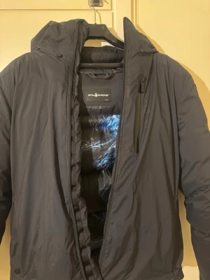 Svart pufferjacka från Sail Racing - Säljer en svart pufferjacka från Sail Racing i storlek M. Jackan har huva, dragkedja framtill och en bröstficka med dragkedja. Insidan har ett coolt tryck och jackan är perfekt för kalla dagar. Passformen är normal och materialet känns slitstarkt och tåligt.