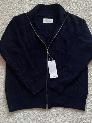 Mörkblå stickad kofta från Maison Margiela - Säljer en mörkblå stickad kofta från Maison Margiela med hög krage och hel dragkedja framtill. Klassisk ribbad struktur och diskreta vita sömmar i nacken. Perfekt för dig som gillar minimalistisk och stilren design.