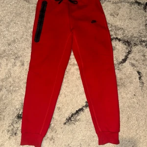 Röda mjukisbyxor från Nike M - Säljer ett par röda mjukisbyxor från Nike i storlek M. Byxorna har resår i midjan, snörning och muddar vid bensluten. En svart dragkedja på ena sidan och Nike-logga framtill. Perfekta för chill eller träning.