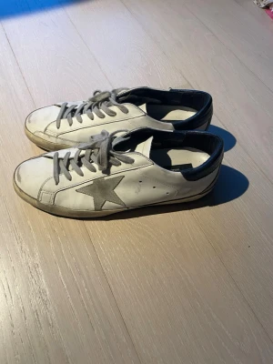Golden Goose Superstar sneakers vit/grå - Nypris är 6000kr. Har kvitto. Golden Goose Superstar sneakers i vitt skinn med grå stjärndetalj på sidan, grå snörning och mörkblå häl. Klassisk låg modell med platt sula och rund tå. Perfekta för dig som gillar en avslappnad men trendig look.