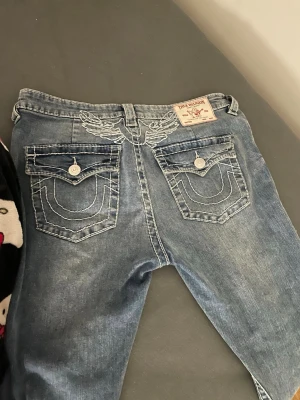 True Religion ljusblå jeans, storlek 31 - Säljer ett par klassiska ljusblå jeans från True Religion med ikoniska kontrastsömmar och broderade detaljer på bakfickorna. Jeansen är boot cut, normal passform och coola ficklock med knappar. Perfekt för dig som gillar streetwear och vill sticka ut. Aldrig använda 💕