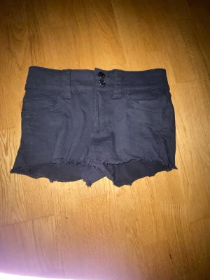 Svarta jeansshorts med rå kant - Säljer ett par svarta shorts i jeansmodell med fransig, rå kant nertill. Shortsen har två knappar framtill, bälteshällor och klassiska fickor både fram och bak. Perfekta till sommaren och ger en cool vibe till din outfit.
