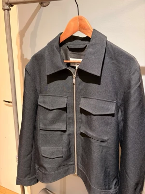 Svart jacka från Maison Margiela - Säljer en svart jacka från Maison Margiela. Jackan har dragkedja, klassisk krage och är tillverkad i ett slätt material som känns lite lyxigt. Perfekt för dig som gillar minimalistisk och clean stil.