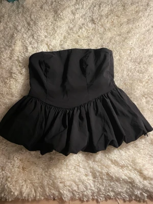 Svart volang tubtopp med dragkedja på sidan  - Säljer en svart axelbandslös tubtopp med volangkant nedtill. Toppen har en bandeauformad överdel, smockad rygg för extra stretch och en diskret dragkedja i sidan. Materialet känns som bomull eller polyester och toppen är croppad i modellen.