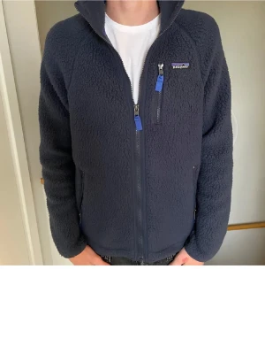 Mörkblå fleecejacka från Patagonia - Mysig mörkblå fleecejacka från Patagonia med hel dragkedja och tre fickor med blå zip-pullers. Jackan har hög krage och en klassisk, lite loose passform. Perfekt för kyliga dagar och enkel att slänga på över en t-shirt.