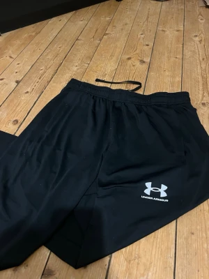 Svarta träningsbyxor från Under Armour - Svarta träningsbyxor från Under Armour med dragsko i midjan och logga på benet. Tillverkade i mjukt och lätt syntetmaterial som andas. Bra passform med raka ben och diskreta fickor på sidorna. Perfekta för gymmet eller chill.