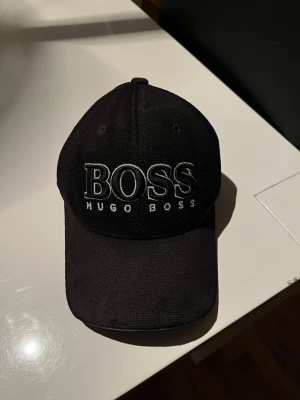 Svart keps från Hugo Boss - Säljer en svart keps från Hugo Boss med tydlig broderad logga framtill. Klassisk böjd skärm och justerbar passform. Tillverkad i ett slitstarkt tyg som ger en sportig och clean look. Säljs inte längre.