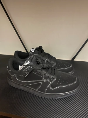 Nike Air Jordan 1 Low svart/vit - Säljer ett par Nike Air Jordan 1 Low i svart med vita kontrastsömmar och broderade detaljer på hälen. Skorna har klassisk snörning, perforerad tåbox och en platt sula. Materialet är troligtvis skinn eller syntet. Ikonisk Air Jordan-logga och bi-detalj bak.