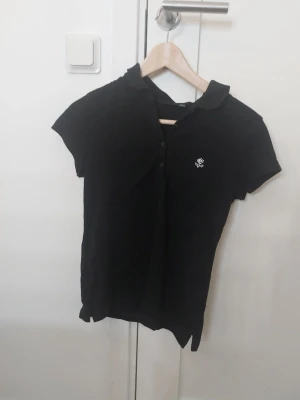 Svart piké från Lindex S - Klassisk svart pikétröja från Lindex i storlek S. Toppen har korta ärmar, krage och knappar framtill samt en liten vit broderad detalj på bröstet. Perfekt till jeans eller kjol för en enkel och clean look.