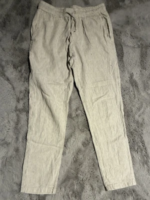 Randiga linnebyxor från ONLY  - Säljer ett par ljusa linnebyxor från ONLY & SONS |med tunna vertikala ränder. Modellen heter Linus Tapered Cropped och har snörning i midjan samt fickor både fram och bak. Perfekta för en avslappnad och stilren look.