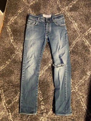 Blå  jeans från Jacob Cohën - Säljer ett par blå slim fit jeans från Jacob Cohën med slitning och hål på ena knät. Jeansen har klassisk femficksdesign, normal midja och är tillverkade i mjukt bomullsmaterial. Snygg tvättad look och diskreta detaljer.