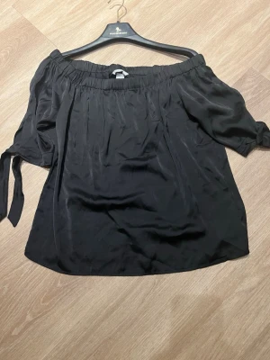 Svart offshoulder blus med knyt - Säljer en svart offshoulder silkes blus med resår upptill och knytband vid ärmsluten. 