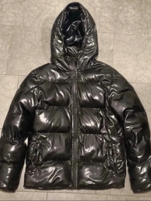 Svart glansig pufferjacka - Säljer en svart pufferjacka med glansig finish och huva. Jackan har dragkedja framtill och breda, vadderade kanaler som ger en riktigt fet look. Perfekt för kalla dagar när du vill hålla stilen och värmen.