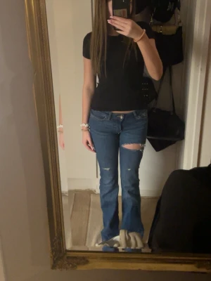 Blå bootcut jeans från only - Säljer ett par blå bootcut jeans med slitna detaljer på låret och knäet. Jeansen har låg midja och klassisk femficksdesign. Perfekta för dig som gillar en avslappnad och trendig look.