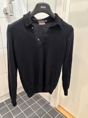 Svart pikétröja i ull från Oscar Jacobson - Klassisk svart pikétröja från Oscar Jacobson i mjuk ull. Tröjan har lång ärm, krage och knappar vid halsen för en clean look. Perfekt till jeans eller chinos för en stilren outfit. Pris kan diskuteras vid snabb affär! 🥂 