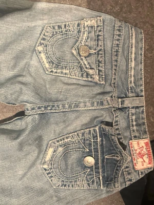 True Religion ljusblå bootcut jeans - Säljer ett par ljusblå jeans från True Religion med ikoniska kontrastsömmar och stora fickor bak. Modellen har låg midja och bootcut-ben, slitningar och hål på ena benet för en extra edgy look. Klassiska metallknappar och detaljerad fickdesign.