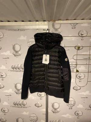 Moncler cardigan - Säljer en svart dunväst från Moncler med huva och ribbstickade ärmar. Västen har dragkedja framtill, två fickor med knapp och klassisk Moncler-logga på ärmen. Materialmix med dun och stickad polyester ger en modern look.
