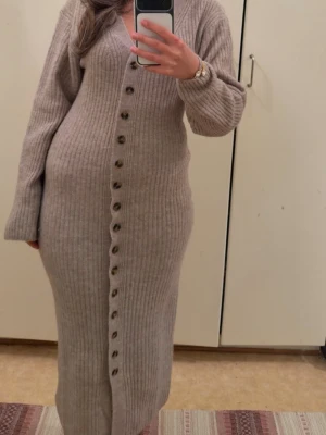 Beige ribbad långklänning med knappar - Mysig beige ribbad långklänning med v-ringning och långa ärmar. Klänningen har en rad dekorativa knappar framtill och är figurnära i modellen. Perfekt för dig som gillar en enkel men ändå stilren look.