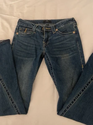 True Religion Joey Low Rise Flare Jeans  - Säljer ett par jättefina  mellanblå True Religion Joey jeans med låg midja och flare-ben. Klassiska kontrastsömmar i gult och snygga ficklock bak med guldiga knappar och broderad logga. Jeansen har fem fickor och är tillverkade i slitstarkt denim. Perfekta för dig som gillar retrovibbar och statement-jeans.💕