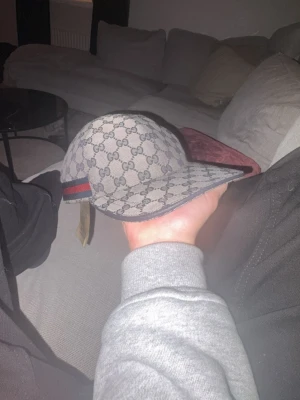 Gucci keps med GG-mönster - Säljer en snygg Gucci keps i beige och marinblått med klassiskt GG-mönster över hela kepsen. Kepsen har justerbart band bak och en röd-blå rand på sidan. Materialet är slitstark bomull och insidan är fodrad i marinblått tyg. Perfekt för dig som vill ha en lyxig och stilren accessoar.