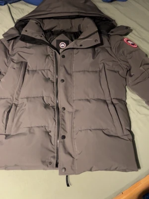 Grå Canada Goose pufferjacka S - Säljer en grå pufferjacka från Canada Goose i storlek S. Jackan har huva, dragkedja och knappar framtill samt två stora fickor. Klassisk patch på ärmen och riktigt skön passform för kalla dagar. Perfekt för dig som vill ha en varm och stilren vinterjacka.                                                Priset är ej hugget i sten!😇😇