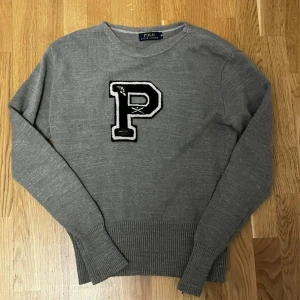 Vintage Polo Ralph Lauren Tröja - Grå stickad tröja från Polo Ralph Lauren med svartvit P-applikation. Klassisk college-stil med rund hals och ribbade muddar. Midjan har krympt något – perfekt för slim fit och passar även Small.