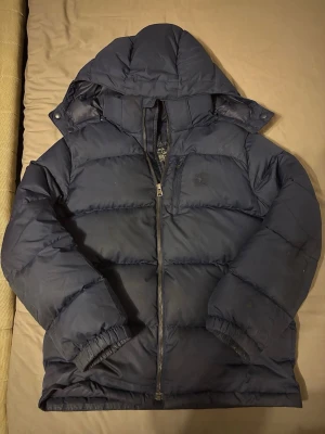 Mörkblå pufferjacka från Ralph Lauren - Säljer en mörkblå pufferjacka från Ralph Lauren. Jackan har huva, dragkedja framtill och en bröstficka med diskret logga. Perfekt för kalla dagar och riktigt skön att bära. Klassisk design och passar till det mesta.