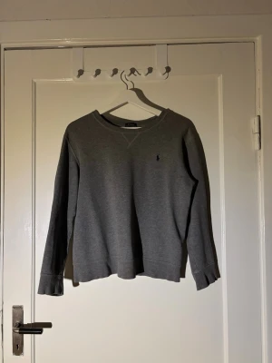Grå sweatshirt från Polo Ralph Lauren - En stilren grå sweatshirt från Polo Ralph Lauren med rund halsringning och diskret broderad logga på bröstet. Tröjan har långa ärmar och är gjord i mjuk bomull, perfekt för chill dagar eller när du vill ha en clean look. Den är i storlek L men är väldigt liten i sin storlek. Jag har normalt S och sitter bra på mig