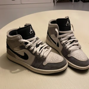 Nike Air Jordan 1 High grå/vit mocka - Nike Air Jordan 1 High sneakers i grå och vit mocka med svarta detaljer och klassisk svart swoosh. Skorna har högt skaft, perforerad tå och vita snören. Yttersulan är svart och ovandelen är tillverkad i en mix av mocka och textil för en clean och stilren look. Har använts drygt en vecka bara.