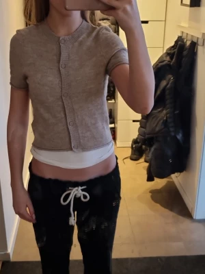 Beige kortärmad kofta från H&M Divided - Kortärmad beige kofta från H&M Divided i storlek XXS. Tröjan har rund hals, ribbade kanter och knappar framtill. Perfekt till lager på lager och enkel att styla med olika outfits.