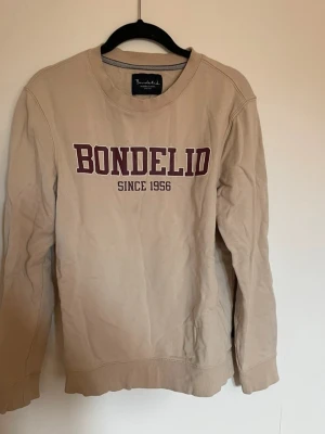 Beige sweatshirt från Bondelid - Säljer en beige sweatshirt från Bondelid med tryck i vinrött framtill. Klassisk rund halsringning och ribbade muddar vid ärmslut och nederkant. Perfekt för en chill och stilren look. Lite liten i storleken.