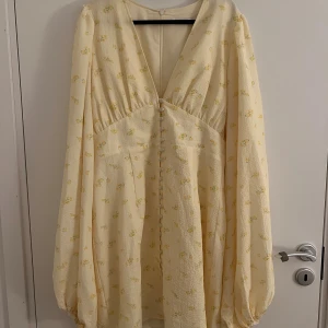 Ljusgul blommig klänning från Adoore - Supersöt ljusgul klänning från Adoore med små gula blommor, v-ringning och klädda knappar framtill. Långärmad med puffiga ärmar och lätt struktur i tyget. Perfekt för sommarens alla tillfällen!